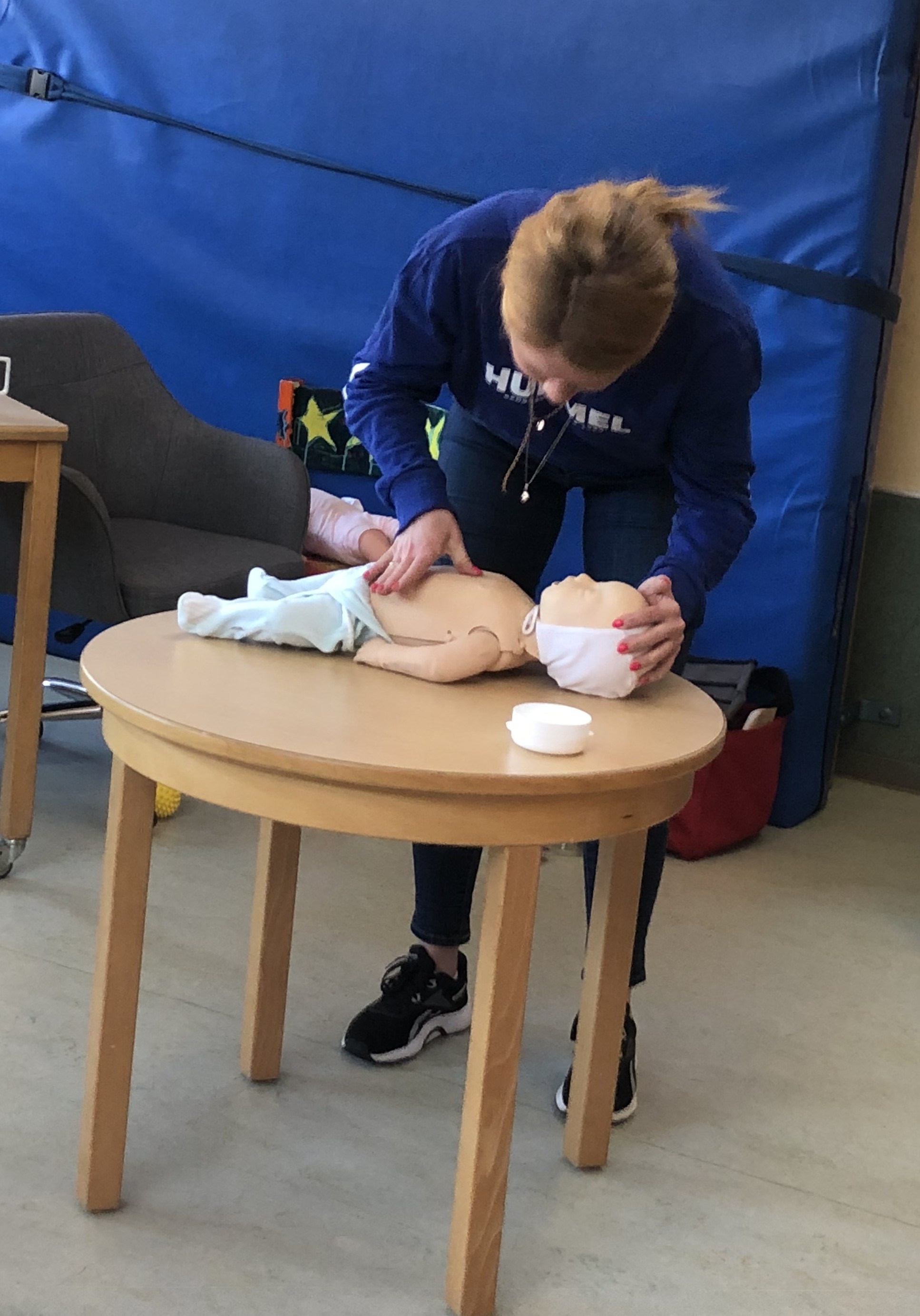 Kursteilnehmerin steht hinter einem Tisch mit einer Reanimationsbabypuppen und hat die Hände auf Kopf und Brust gelegt.